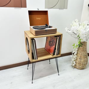 May include: A light wood record player stand with four thin black metal hairpin legs and a square open storage compartment containing vinyl records. A portable record player with a tan exterior and the word Vintage is placed on top.