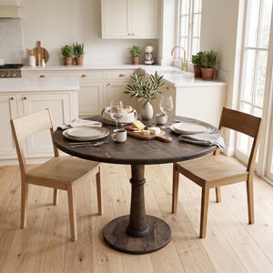 Op de afbeelding: Een ronde, donkerbruine houten eettafel met een centrale poot, gecombineerd met twee lichtgekleurde houten stoelen. De tafel is gedekt met witte keramische borden, glazen, een houten serveerplank met kaas en een vaas met groene takken.