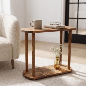 Puede incluir: Una mesa auxiliar de madera con una superficie y base ovaladas y lisas, conectadas por cuatro patas cilíndricas. La mesa sostiene una taza de cerámica beige, un libro cerrado con gafas encima y un pequeño jarrón de cristal con flores.