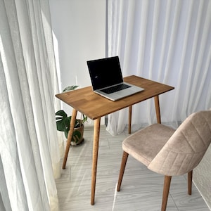 Puede incluir: Un pequeño escritorio de madera con un portátil encima, una silla beige y una planta en maceta. El escritorio tiene cuatro patas cónicas y una tapa rectangular. La silla tiene asiento y respaldo acolchados. Cortinas blancas transparentes en el fondo.