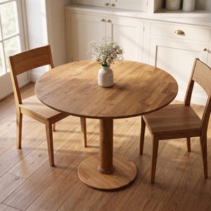 May include: A round wooden dining table with a central pedestal base and two matching wooden chairs. A small white ceramic vase containing a bouquet of white flowers sits in the center of the table, which features a natural wood grain finish.