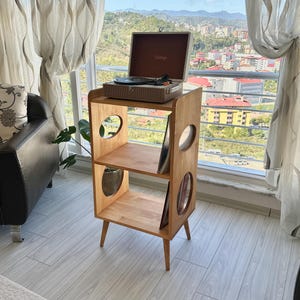 May include: A light brown wooden record player stand with three tiers and circular cutouts on the side panels. The stand features four tapered legs and holds a portable suitcase-style record player on the top shelf, with vinyl records stored vertically on the lower shelves.