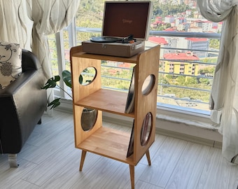 May include: A light brown wooden record player stand with three tiers and circular cutouts on the side panels. The stand features four tapered legs and holds a portable suitcase-style record player on the top shelf, with vinyl records stored vertically on the lower shelves.
