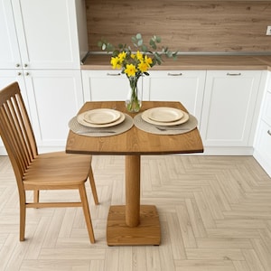 Puede incluir: Una mesa de comedor de madera con una parte superior cuadrada y una base de pedestal única. Dos cubiertos con platos y cubiertos están colocados sobre la mesa. Una silla de madera está a la izquierda. Un jarrón con flores amarillas está sobre la mesa.