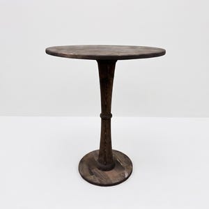 Op de afbeelding: Een ronde, donkerbruine houten tafel met een glad, cirkelvormig blad dat wordt ondersteund door een enkele centrale kolom die naar het midden toe taps toeloopt en aan de basis uitloopt. De houtnerf is overal zichtbaar.