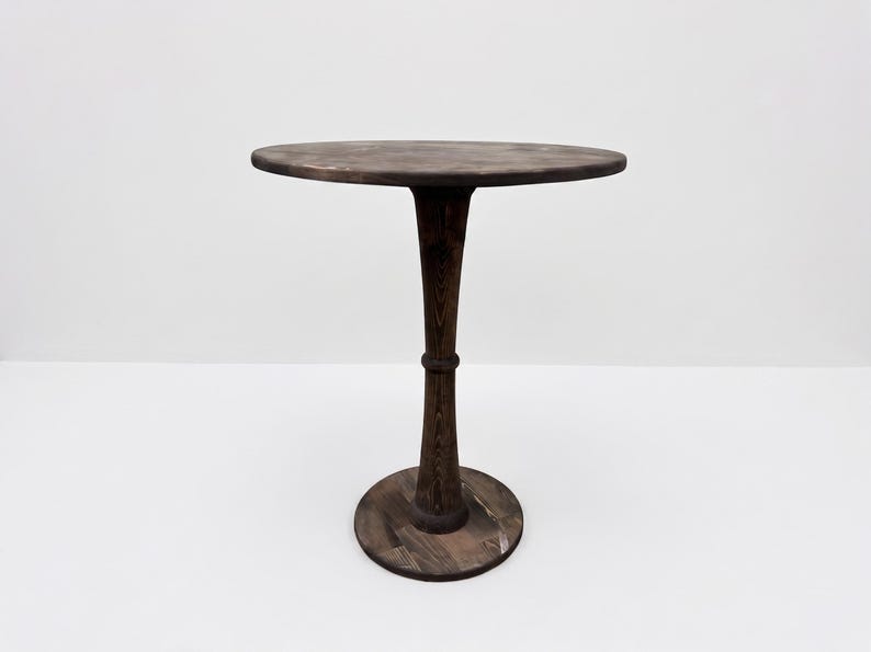 Op de afbeelding: Een ronde, donkerbruine houten tafel met een glad, cirkelvormig blad dat wordt ondersteund door een enkele centrale kolom die naar het midden toe taps toeloopt en aan de basis uitloopt. De houtnerf is overal zichtbaar.
