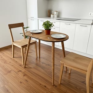 Op de afbeelding: Een kleine, ovale houten eettafel met twee bijpassende houten stoelen. De tafel heeft twee borden en een kleine plant in een pot. De stoelen en tafel zijn gemaakt van lichtgekleurd hout. De tafel is ongeveer 100 cm lang.