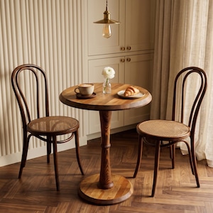 May include: A round wooden pedestal table with a dark brown finish, accompanied by two matching bentwood chairs featuring woven cane seats. On the table sits a ceramic mug with a beverage, a small glass vase with a white flower, and a plate with a croissant.