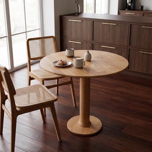 Pode incluir: Uma mesa de jantar redonda de madeira com uma base de pedestal, acompanhada por duas cadeiras com encostos e assentos de vime tecido. A mesa está posta com duas canecas, um prato pequeno com comida e um vaso pequeno. A sala tem piso e armários de madeira escura.