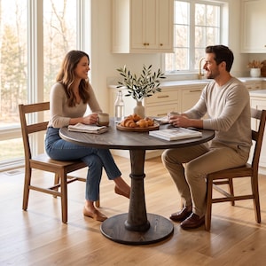 Op de afbeelding: Een ronde eettafel van donker hout met een centrale kolomvoet, vergezeld door twee houten stoelen. Het tafeloppervlak bevat een houten serveerblad met gebak, een doorzichtige glazen waterfles en een vaas met groene bladeren.