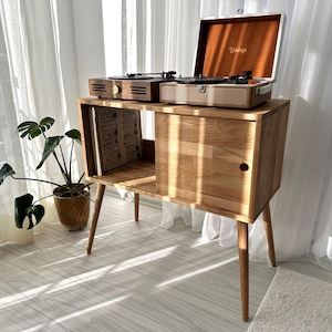 May include: Wooden record player stand with a mid-century modern design. The stand has a light brown finish, with a cabinet and open shelf for vinyl records. Two record players are on top, one in a brown case and the other in a tan case.