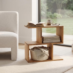 Può includere: Un tavolino in legno color marrone chiaro con un design a forma di S dotato di due scomparti aperti. Il piano superiore ospita un libro aperto, una tazza in ceramica e degli occhiali, mentre lo scomparto inferiore contiene un cesto intrecciato.
