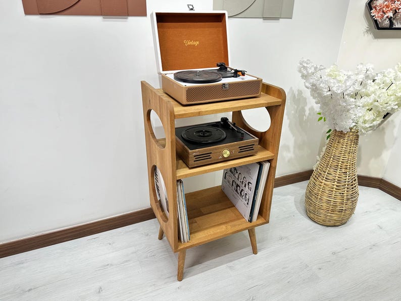 May include: A wooden record player stand with three shelves. The top shelf has a white record player with a brown lid and a turntable. The middle shelf has a black record player with a brown lid. The bottom shelf has two vinyl records. The stand has a rounded cutout on each side.