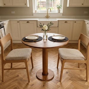 May include: A round wooden dining table with two chairs in a kitchen setting. The table is set with two plates on black placemats. The chairs have light-colored upholstered seats. A vase of flowers sits in the center of the table.
