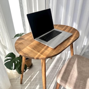 Puede incluir: Un portátil plateado con pantalla negra está sobre una pequeña mesa ovalada de madera. La mesa tiene tres patas y un acabado de madera natural. Una planta en maceta con hojas verdes grandes está al lado de la mesa. La escena está iluminada por la luz del sol.