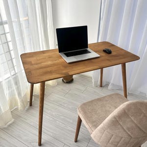 Puede incluir: Un escritorio de madera con un portátil y un ratón. El escritorio tiene un acabado marrón claro y esquinas redondeadas. Una silla beige está frente al escritorio. La habitación tiene cortinas blancas y una ventana. El escritorio mide unos 120 cm de largo.