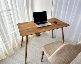 May include: A wooden desk with a laptop and mouse on top. The desk has a light brown finish and rounded corners. A beige chair is positioned in front of the desk. The room has white curtains and a window.