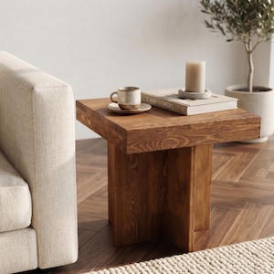 May include: A square wooden side table with a thick top and a cross-shaped base. The table is made of medium-brown wood with a visible grain pattern. It features a smooth, flat surface and a sturdy, geometric construction that supports the tabletop.