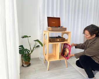 May include: A wooden record player stand with two turntables and a record. The stand has a mid-century modern design with circular cutouts. A woman is holding a record with a pink cover.