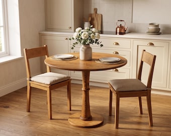 Op de afbeelding: Een ronde houten eettafel met twee bijpassende stoelen. De tafel heeft een voetstuk en is gedekt met borden en placemats. Een vaas met witte bloemen staat in het midden. De stoelen hebben gewatteerde zittingen.