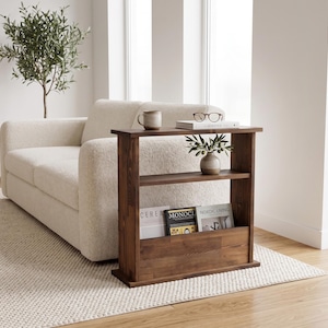 May include: A rectangular dark brown wooden console table featuring two open shelves and a lower storage compartment. The top surface holds a ceramic mug, a pair of glasses, and a book. The middle shelf holds a small vase, while the bottom section displays three magazines titled Cereal, Monocle, and Nordic Living.