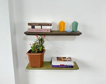 Può includere: Due mensole galleggianti in legno, una marrone scuro e una verde, sono montate su una parete bianca. La mensola superiore contiene libri e candele. La mensola inferiore contiene libri e una pianta in vaso. Le mensole sono una soluzione di archiviazione decorativa.