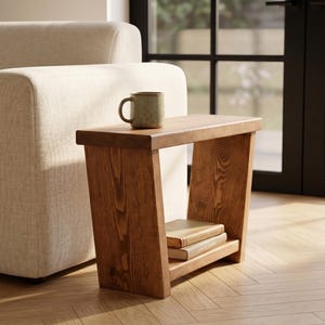 May include: A rectangular wooden side table with a natural brown wood grain finish, featuring a flat top surface and a lower shelf. A ceramic mug sits on the top surface, and two books are stacked on the lower shelf of the wooden table.