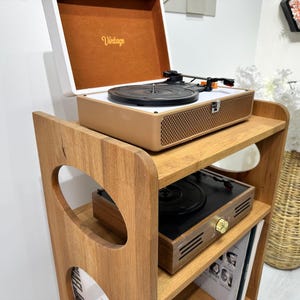 May include: A wooden record player stand with three shelves. The top shelf has a vintage record player with a brown and white exterior. The middle shelf has a black record player with a brown wooden base. The bottom shelf has a stack of vinyl records.