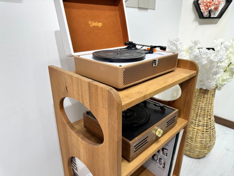 May include: A wooden record player stand with three shelves. The top shelf has a vintage record player with a brown and white exterior. The middle shelf has a black record player with a brown wooden base. The bottom shelf has a stack of vinyl records.