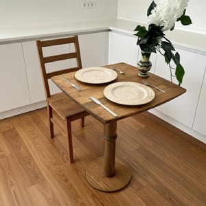 Puede incluir: Una pequeña mesa cuadrada de madera con una sola base de pedestal, preparada para dos personas. Dos platos decorativos, cubiertos y una silla de madera están colocados sobre la mesa. Un jarrón con flores blancas está cerca. La mesa mide unos 76 cm cuadrados.