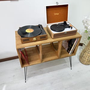 Puede incluir: Un soporte de madera para tocadiscos con tres estantes para guardar discos de vinilo. El soporte tiene patas de horquilla de metal negro y presenta dos tocadiscos en la parte superior. Uno de los tocadiscos es blanco con una tapa marr&oacute;n y el otro es negro con una tapa transparente.