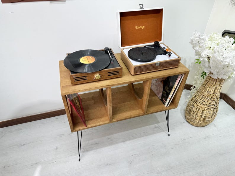 Puede incluir: Un soporte de madera para tocadiscos con tres estantes para guardar discos de vinilo. El soporte tiene patas de horquilla de metal negro y presenta dos tocadiscos en la parte superior. Uno de los tocadiscos es blanco con una tapa marr&oacute;n y el otro es negro con una tapa transparente.