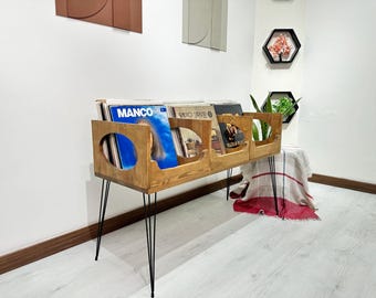 Könnte beinhalten: Ein Holz-Schallplattenständer mit schwarzen Metallbeinen. Der Ständer hat drei Regale und ist mit Schallplatten gefüllt. Der Ständer steht in einem Raum mit weißen Wänden und einem Holzboden.