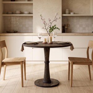 Op de afbeelding: Een ronde eettafel van donkerbruin hout met een centrale kolomvoet, vergezeld door twee lichtgekleurde houten stoelen. De tafel is gedekt met twee borden, twee wijnglazen, twee stompkaarsen en een vaas met bloeiende takken.