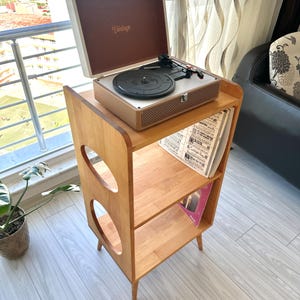 May include: A light brown wooden record player stand with two open shelves and circular side cutouts. A portable suitcase-style record player with a white exterior and brown interior featuring the word Vintage is placed on the top surface.