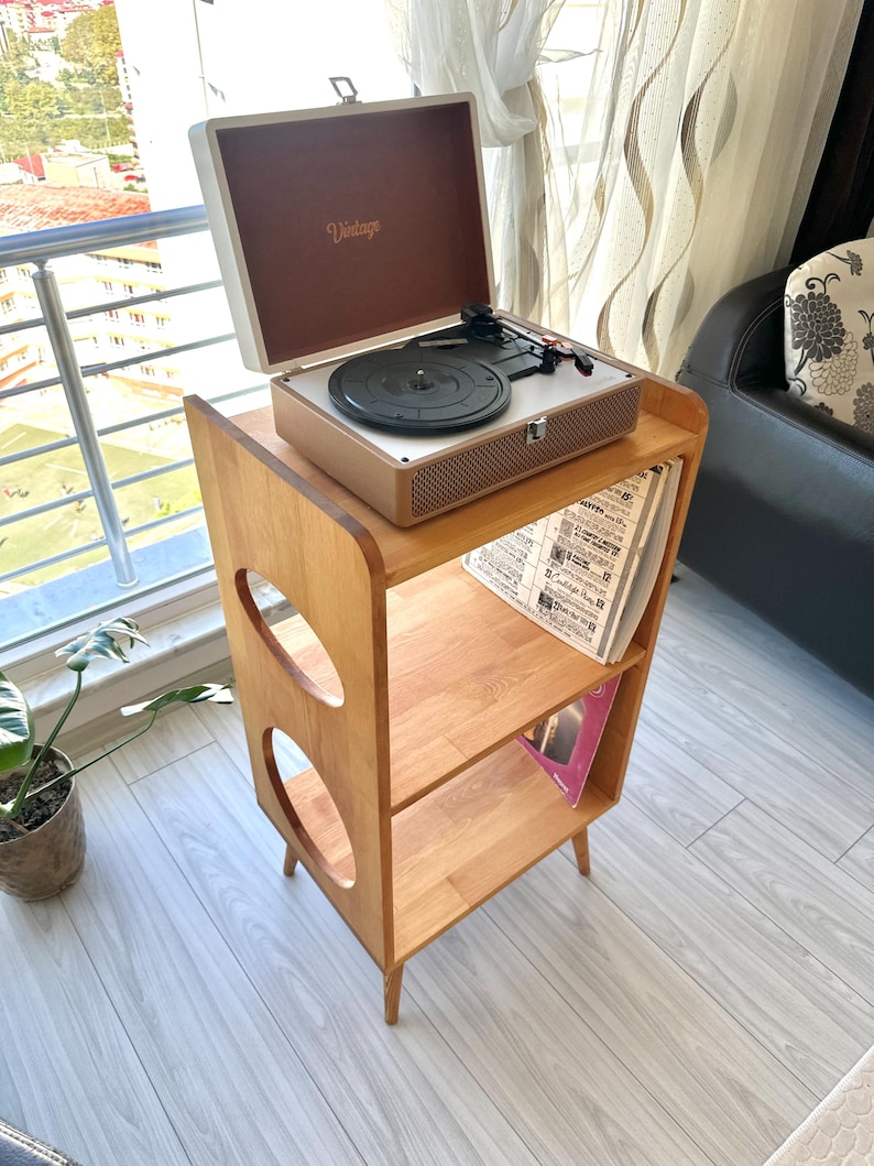 May include: A light brown wooden record player stand with two open shelves and circular side cutouts. A portable suitcase-style record player with a white exterior and brown interior featuring the word Vintage is placed on the top surface.