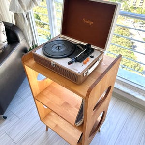 May include: A portable suitcase-style record player with a cream and brown exterior, featuring the word Vintage on the inner lid and umie on the control panel, resting on a light brown wooden stand with three open shelves and circular side cutouts.