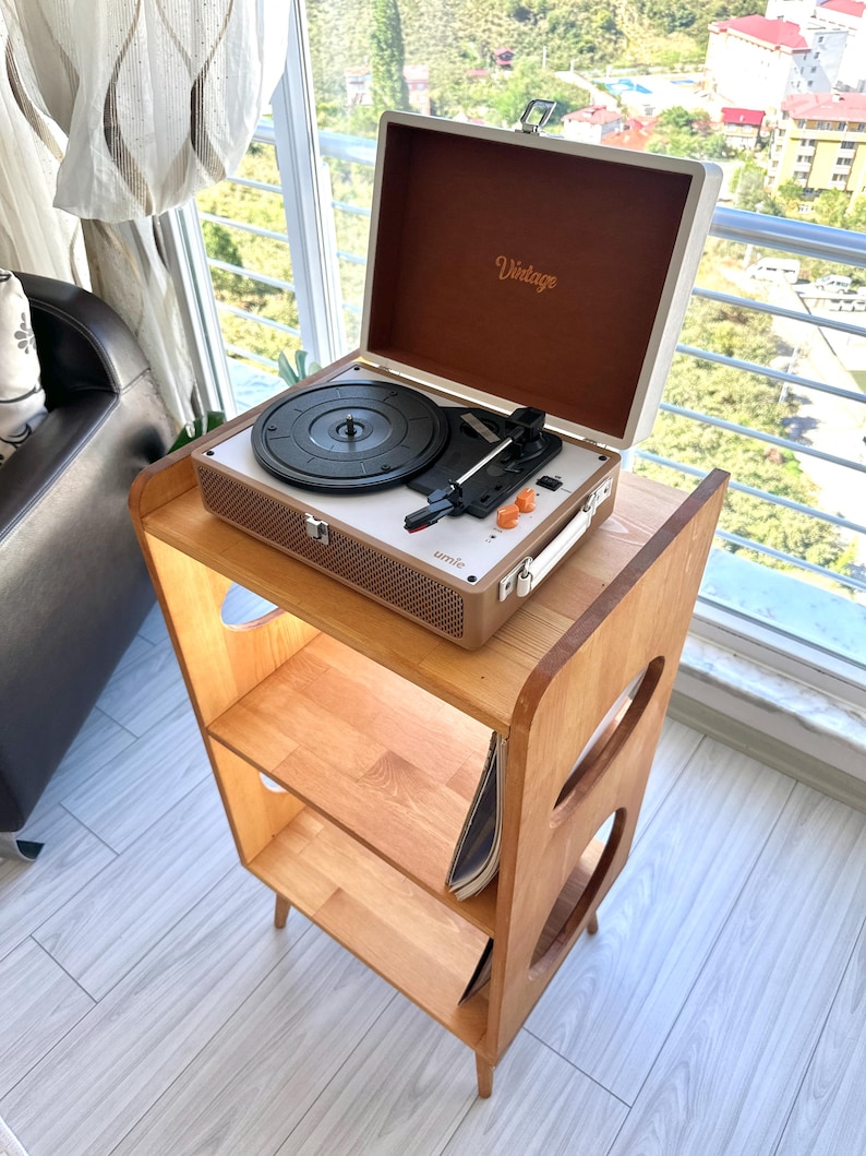 May include: A portable suitcase-style record player with a cream and brown exterior, featuring the word Vintage on the inner lid and umie on the control panel, resting on a light brown wooden stand with three open shelves and circular side cutouts.