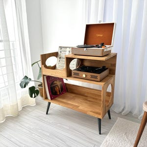 May include: Wooden record player stand with two levels, holding a vintage-style record player with a tan exterior and a brown interior. The stand has black legs and a circular cutout. A record album is visible on the lower shelf.