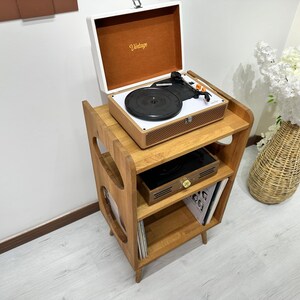 May include: A wooden record player stand with three shelves. The top shelf has a vintage record player with a white lid and a black turntable. The middle shelf has a small wooden box and the bottom shelf has vinyl records.
