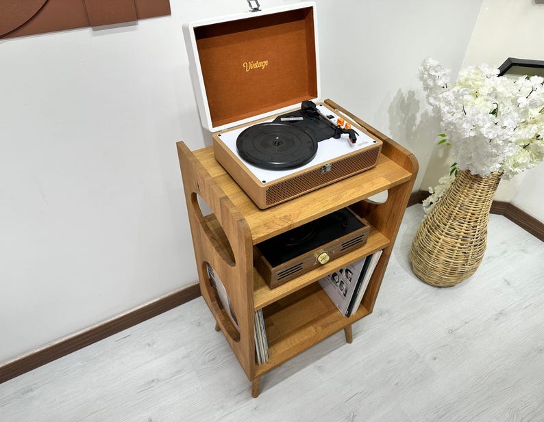 May include: A wooden record player stand with three shelves. The top shelf has a vintage record player with a white lid and a black turntable. The middle shelf has a small wooden box and the bottom shelf has vinyl records.