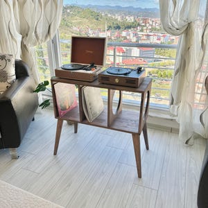 May include: A wooden record player stand with two turntables. One turntable is in a brown case with the word "Vintage" on it. The other turntable is brown. The stand has a shelf for record storage. The stand is in front of a window with a view of a city.