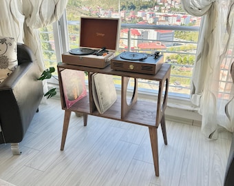 May include: A wooden record player stand with four tapered legs and two open storage compartments containing vinyl records. Two portable record players are placed on the top surface, one with a cream-colored lid and one with a dark wood finish.