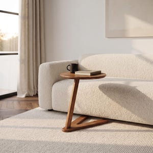 May include: A round wooden side table with a single angled support leg and a V-shaped base. The table surface holds a black mug and a closed book. The furniture is constructed from a medium-toned wood with a visible grain, featuring a smooth finish.