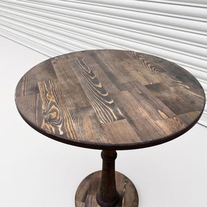 Op de afbeelding: Een ronde houten tafel met een donkerbruine gebeitste afwerking die een zichtbaar houtnerfpatroon vertoont. Het tafelblad is opgebouwd uit meerdere samengevoegde planken en rust op een centrale kolomvoet met een ronde voetplaat.