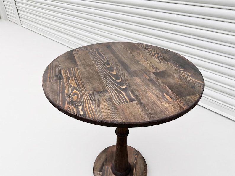 Op de afbeelding: Een ronde houten tafel met een donkerbruine gebeitste afwerking die een zichtbaar houtnerfpatroon vertoont. Het tafelblad is opgebouwd uit meerdere samengevoegde planken en rust op een centrale kolomvoet met een ronde voetplaat.