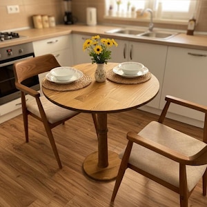 Może przedstawiać: Mały, okrągły drewniany stół jadalny z dwoma pasującymi krzesłami. Na stole znajdują się dwie białe miski na tkanych podkładkach i wazon z żółtymi kwiatami. Krzesła mają jasne, tapicerowane siedziska i drewniane ramy.