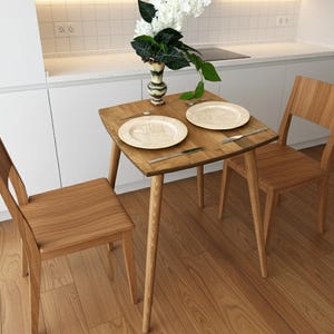 Op de afbeelding: Een kleine, vierkante houten eettafel met twee bijpassende stoelen. De tafel is gedekt met twee borden, bestek en een vaas met bloemen. De tafel is ongeveer 76 cm breed en 76 cm lang.