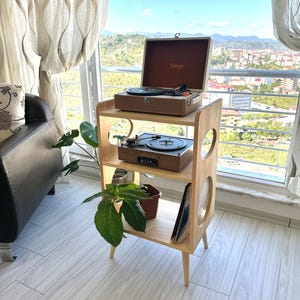 Puede incluir: Un soporte de tocadiscos de madera clara con dos tocadiscos y un estuche de transporte marrón. El soporte tiene tres estantes, con una planta y discos en los estantes inferiores. El soporte está frente a una ventana con vista a la ciudad.