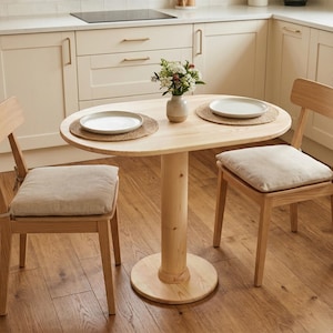 May include: A light wood oval dining table with two matching chairs. The table has a single central support and a round base. Each chair has a light-colored cushion. Two white plates sit on the table, each on a woven placemat. A small vase of flowers is in the center.
